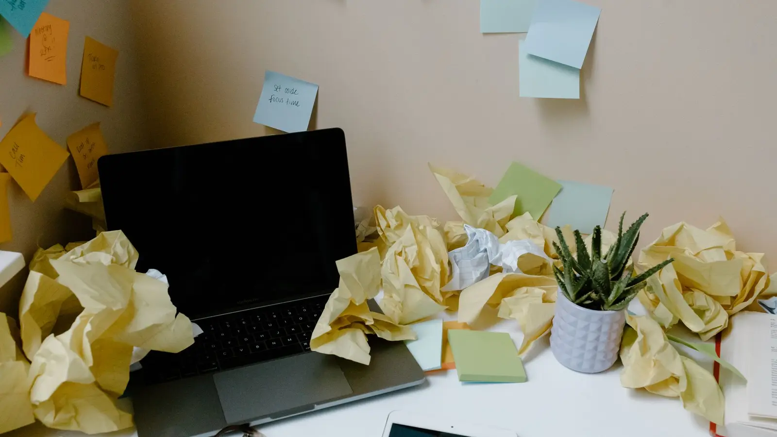 Meja kerja dengan laptop, sticky note di dinding, dan kertas kuning yang diremas